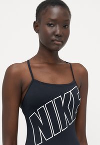 Svart sportstopp med tynne stropper, med en stor hvit Nike-logo over fronten. Glatt, elastisk stoff, figurnær design.