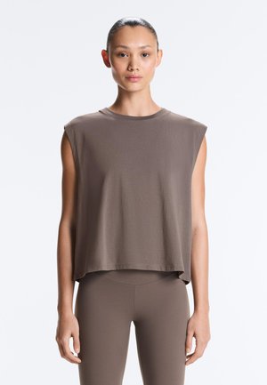 Kvinde med mørkt hår samlet bagpå iført ærmeløs, løs brun top og matchende brune tætsiddende bukser, stående mod en ensartet baggrund.