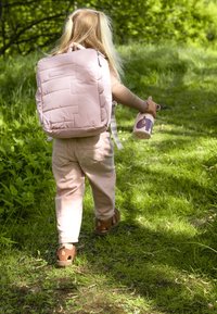 Vaaleanpunainen pehmustettu reppu säädettävillä remmeillä, lapsen selässä, joka kävelee nurmikolla ja pitää violettia eläinaiheista mukia.