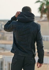 Schwarze Lederjacke mit abnehmbarer grauer Kapuze, mit gesteppten Schulterakzenten und verstellbaren Seitenriemen. Strukturierter Finish.