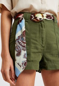 Pantaloni verdi con vita alta, dotati di una fibbia circolare marrone. Una sciarpa decorata in blu chiaro, marrone e crema è annodata in vita.