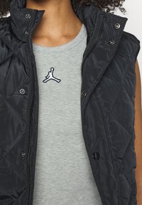 Black quilted vest with a high collar and snap buttons, layered over a light grey T-shirt featuring a black embroidered Jumpman logo.