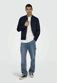 Marinblå bomberjacka med dragkedja, bärs över en vit t-shirt. Ljusblå denimjeans och svarta sneakers fulländar outfiten.