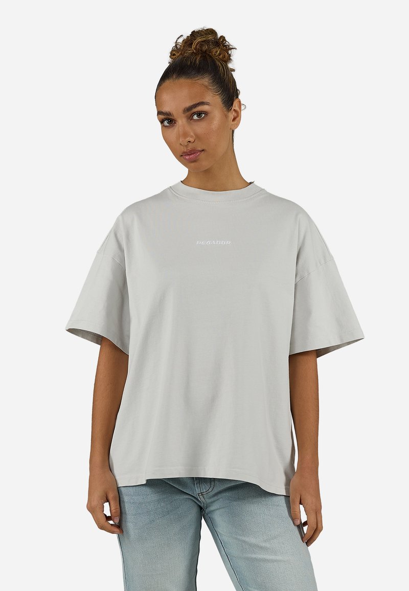 Γυναίκα με ρίγες μαλλιά σε κότσο, φοράει ένα φαρδύ ανοιχτό γκρι μπλουζάκι και ανοιχτό μπλε τζιν, στέκεται μπροστά σε μια απλή λευκή φόντο.