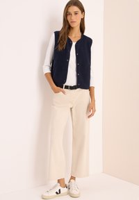 Marine fleece vest met drukknoopjes, gedragen over een wit shirt. Gecombineerd met beige wijde broek en witte sneakers met zwarte accenten.