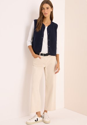 Chaleco de forro polar azul marino con botones a presión, usado sobre una camisa blanca. Combinado con pantalones de pierna ancha beige y zapatillas blancas con detalles en negro.