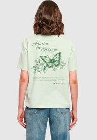 Lys grøn bomulds t-shirt med et sommerfugle grafisk design og blomsterdesign på ryggen. Teksten lyder "Flutter & Bloom" og "Nature's Touch."