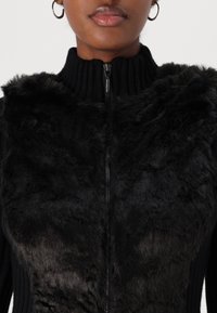 Nærbillede af en person iført en sort sweater med lynlås, der har en struktureret, blød plys frontpanel og ribbet krave.