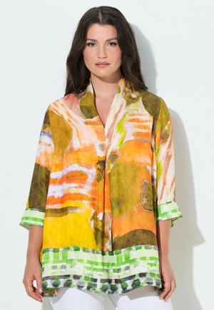 Kobieta nosząca luźną, kolorową bluzkę z abstrakcyjnymi pomarańczowymi, żółtymi, zielonymi i białymi wzorami oraz rękawami do łokcia, stojąca przy jednolitej ścianie.