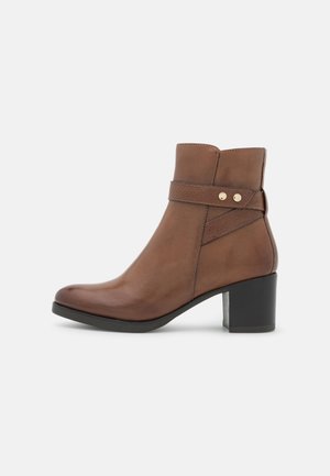 Bottine en cuir marron avec bout arrondi, petit talon carré, et une sangle latérale ornée de deux boutons dorés. Texture lisse, design minimaliste.