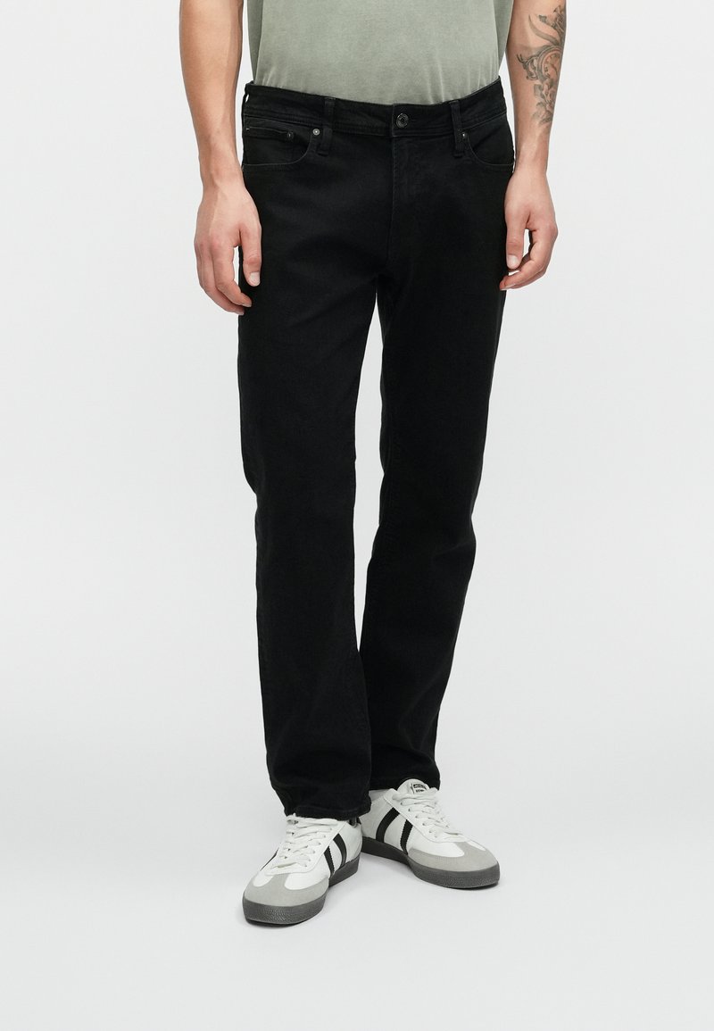 Vaqueros de mezclilla negra con un corte recto, que cuentan con un diseño clásico de cinco bolsillos y una cintura con botones, combinados con zapatillas gris y blancas.