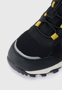 Melnas un pelēkas sporta kurpes ar sietu augšdaļu, pastiprinātu purngali, dzelteniem auklu cilpiņām un teksturētu zoli uz baltā fona.