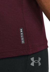 Camisa deportiva granate con textura suave, que presenta una etiqueta negra con la inscripción "RUSH" y un logo de Under Armour en gris sobre pantalones negros.