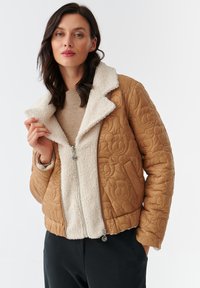 Gequilted beige jack met een fluffy witte kraag en voering. Voorzien van een ritsluiting en een berenpatroon op het buitenmateriaal.