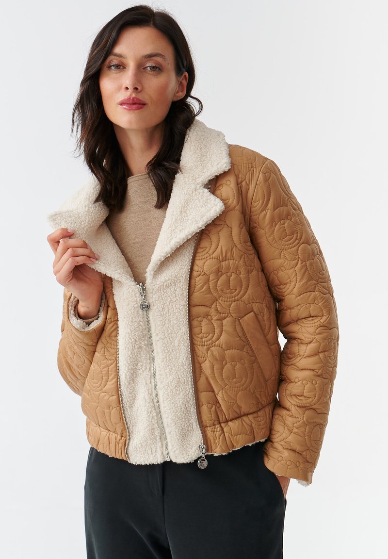Gequilted beige jack met een fluffy witte kraag en voering. Voorzien van een ritsluiting en een berenpatroon op het buitenmateriaal.