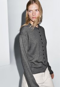 Cardigan gris à manches longues avec un devant à boutons. Présente une texture douce et une coupe décontractée, avec des boutons noirs et un détail de col.