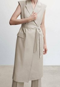 Beżowa sukienka-płaszcz bez rękawów z paskiem w talii, dwoma przednimi kieszeniami, kołnierzem z klapami i teksturowaną tkaniną. Gładki, dopasowany krój.
