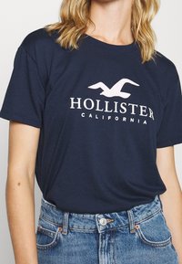 Camiseta de algodón azul marino con el logo blanco "HOLLISTER" y un gráfico de gaviota, combinada con pantalones cortos de mezclilla de cintura alta.