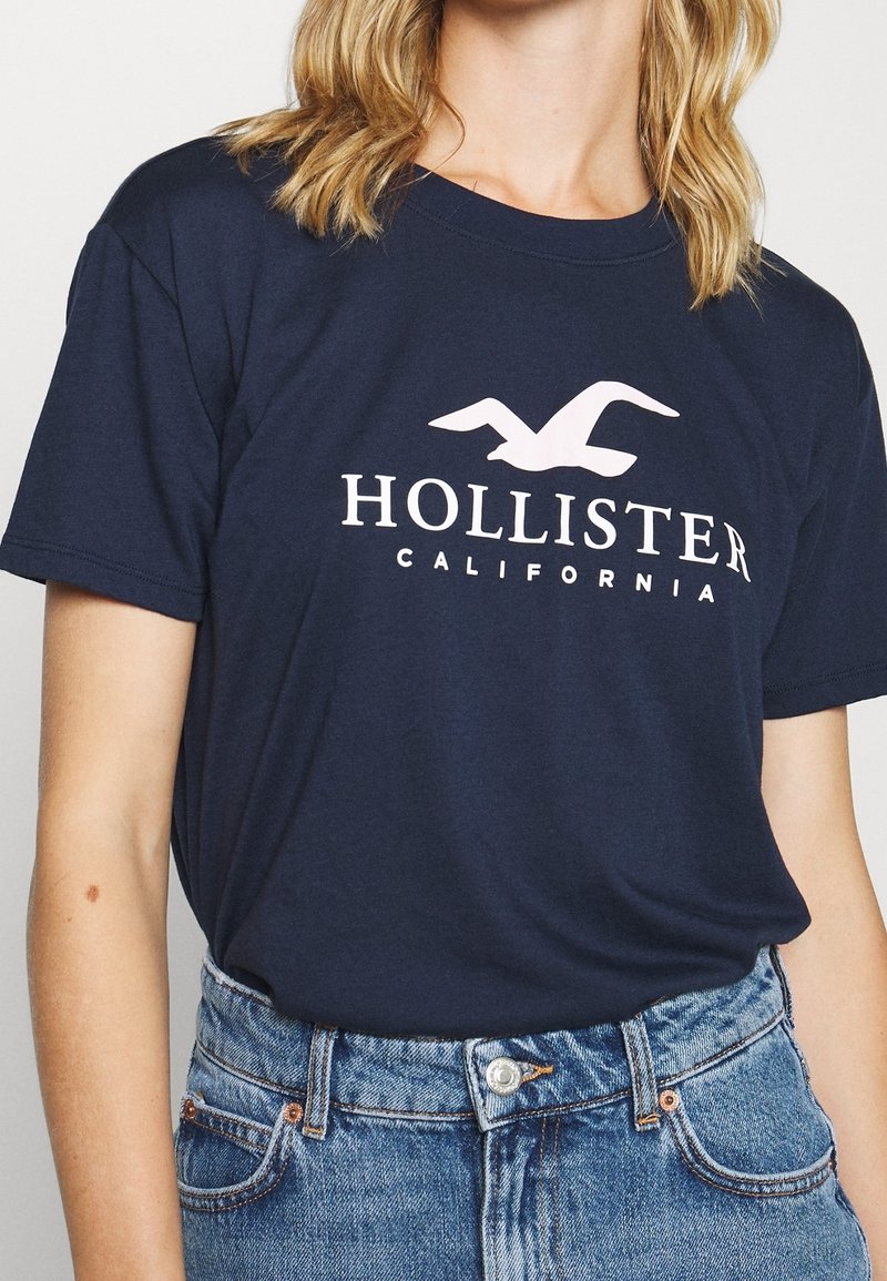 Camiseta de algodón azul marino con el logo blanco "HOLLISTER" y un gráfico de gaviota, combinada con pantalones cortos de mezclilla de cintura alta.