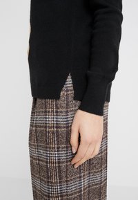 Person trägt einen schwarzen langärmeligen Pullover mit seitlichem Schlitz und einen braunen karierten Rock, die Hand ruht auf der Seite des Rocks.