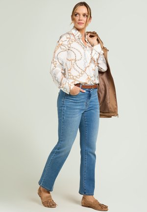 Femme portant un chemisier blanc à motif, un jean bleu avec une ceinture tressée marron, tenant une veste beige sur l'épaule, debout devant un fond uni.