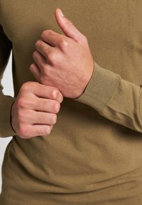 Olivově zelená košile s dlouhým rukávem a žebrovanými manžetami. Detailní záběr na ruce držící látku, ukazující hladkou texturu a přiléhavý střih.