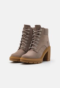 Brown suede ankle boots with a high block heel, featuring metal eyelets and gray laces, textured sole, and logo detail on the side.