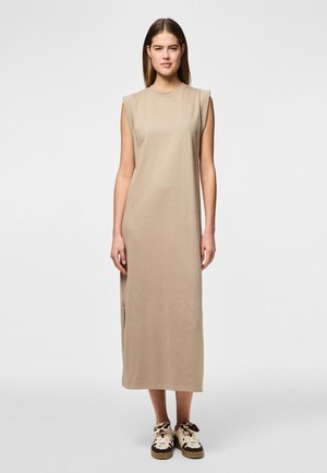 Robe sans manches beige avec une coupe droite, longueur genoux, fabriquée en tissu doux. Présente une fente latérale et un col rond. Portée avec des baskets à motifs.