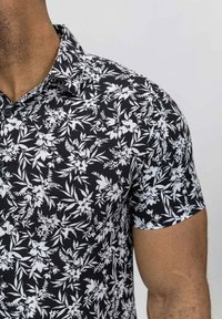 Chemise noire à manches courtes avec un motif floral blanc. Fabriquée en tissu léger, avec un col à boutons et une coupe décontractée.