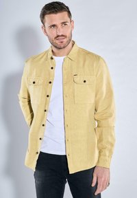 Hellgelbe, knöpfbare Hemd aus strukturiertem Stoff mit zwei Brusttaschen und dunklen Knöpfen, getragen über einem weißen T-Shirt.