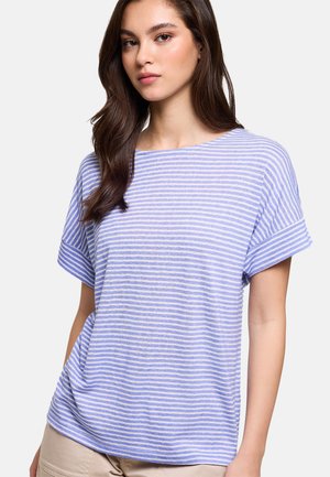 Joven con cabello largo y castaño, vestida con una camiseta de manga corta a rayas morado claro y blanco, y pantalones beige, de pie frente a un fondo liso.