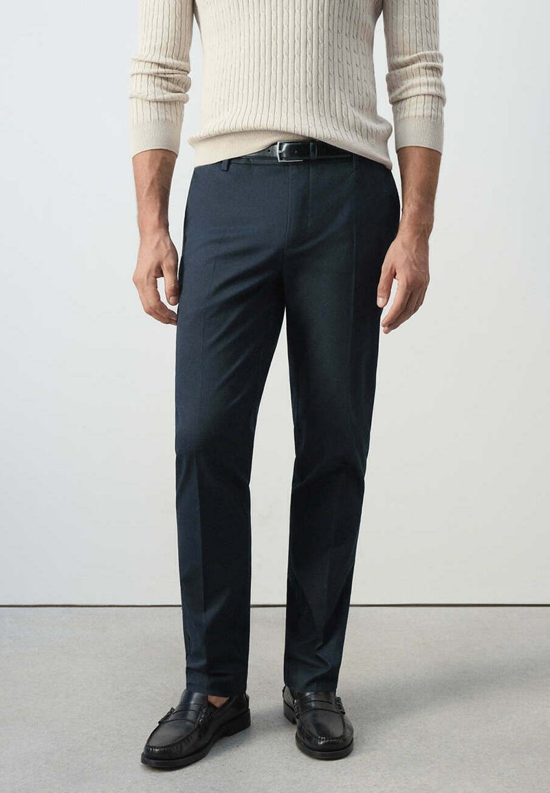 Homme portant un pull côtelé beige, une ceinture noire, un pantalon de costume bleu marine foncé et des mocassins en cuir noirs, debout sur un sol gris.