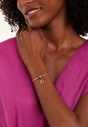 Silver bracelet with a textured band and a charm shaped like a letter. Worn on a hand against a vibrant pink fabric background.