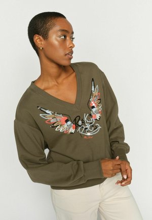 Nainen pukeutuneena oliivinvihreään V-kaulaiseen collegepaitaan, jossa on paljeteilla koristeltu siipikuvio ja "Rock & Roll" -teksti, yhdistettynä vaalean beigeihin housuihin.