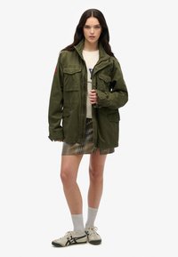 Giacca utilitaria verde oliva con quattro tasche frontali, chiusura a bottoni e vestibilità rilassata, abbinata a una gonna a quadri e sneakers.