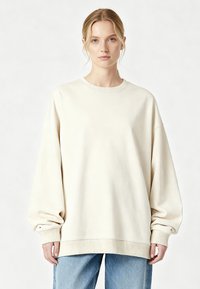 Gaismas bēša oversize džemperis ar ribotu aproču un apakšmalas malu. Mīksta tekstūra un nolaistu plecu dizains. Kombinēts ar gaiši ziliem džinsiem.