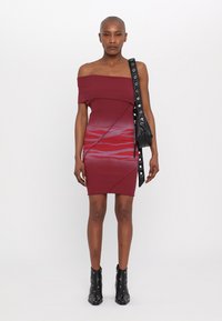 Abito fitted bordeaux con spalle scoperte e motivi ondulati a gradiente, abbinato a una borsa nera con dettagli in argento e stivali neri alla caviglia.