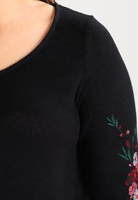 Haut en maille noire avec un col rond en forme de scoop et une broderie florale rouge et verte sur la manche droite. Texture douce et coupe ajustée.