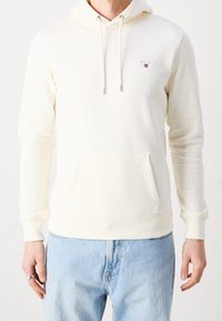 Sudadera color crema de tejido suave con un bolsillo frontal, capucha ajustable con cordón, y logo bordado en el pecho. Presenta puños acanalados.