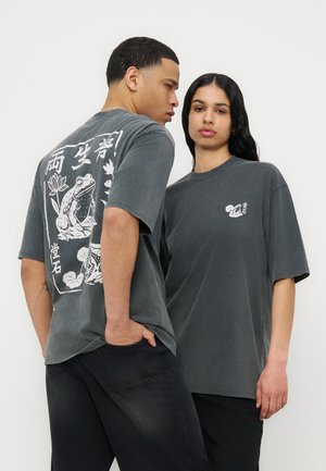 Δύο μοντέλα φορούν υπερμεγέθη σκούρα γκρι μπλουζάκια με λευκά γραφικά βάτραχου και λουλουδιών· το ένα κοιτάζει προς τα εμπρός, ενώ το άλλο δείχνει το μεγάλο σχέδιο στην πλάτη.