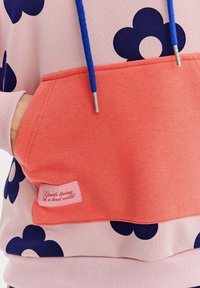 Mikina s kapucňou vyrobená z ružovej látky s námorníckymi kvetinovými vzormi. Má koralovú kapsu s ružovou etikettou, na ktorej je biely text. Modré šnúrky.