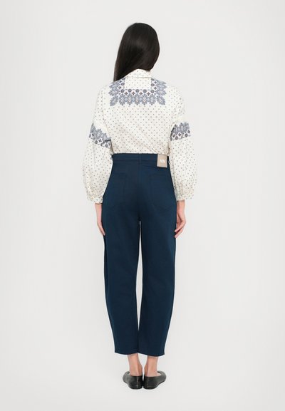 Blusa branca com padrões azuis e mangas bufantes, combinada com calças navy de cintura alta. Sapatos planos pretos completam o visual.