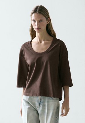 Frau mit langen Haaren trägt ein locker sitzendes braunes Shirt mit Rundhalsausschnitt und halblangen Ärmeln sowie hellblaue Jeans vor einfachem Hintergrund.