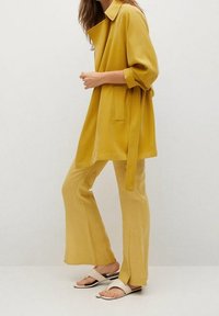 Manteau jaune surdimensionné avec col et taille ceinturée ; associé à un pantalon évasé jaune assorti et des sandales blanches. Texture lisse.