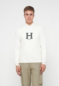 Felpa bianca con colletto rotondo, dotata di un logo "H" grigio scuro e "Hackett" sul petto. Abbinata a pantaloni cargo beige chiaro.