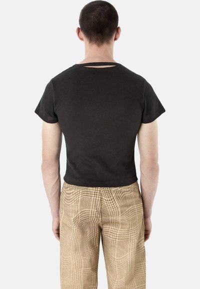 Homme de dos portant un t-shirt noir court et un pantalon beige à carreaux, sur un fond clair uni.