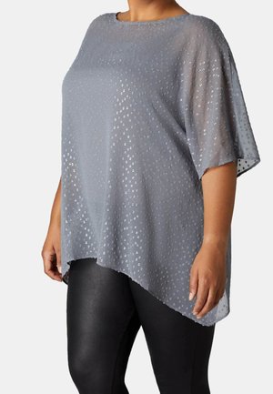 Průsvitný šedý top s krátkými rukávy, zdobený puntíkovaným vzorem a nerovným lemem. Kombinován s černými legínami.