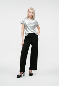 Kvinde med platinblond hår iført en sølvfarvet kortærmet top, sorte vide bukser og sorte højhælede sko med sløjfer.