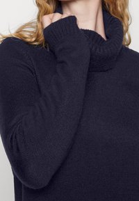 Pull en maille bleu marine avec un col roulé haut, des poignets côtelés et une finition texturée. Tissu doux et extensible avec un aspect confortable.