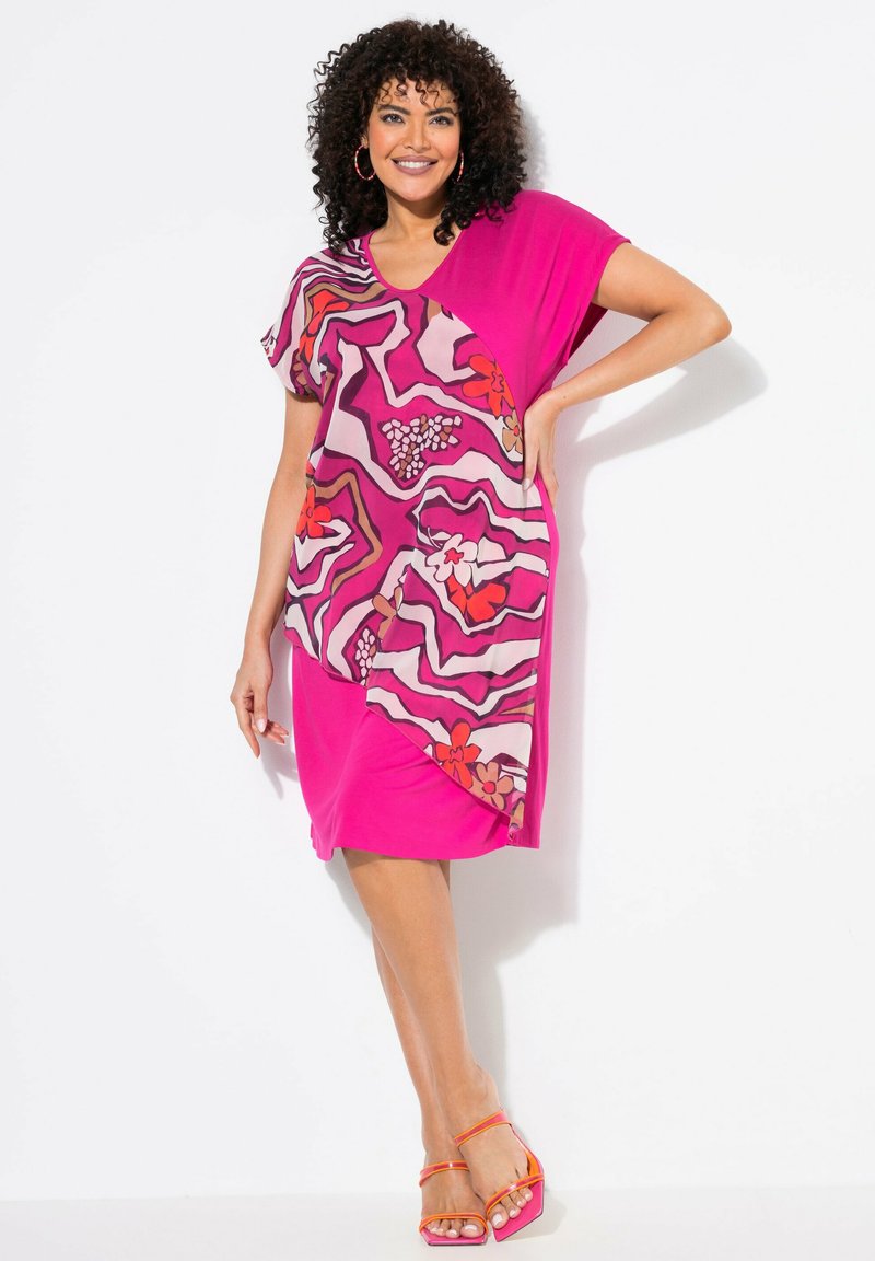 Vestido rosa con un diseño fluido, adornado con patrones florales abstractos y una tela contrastante en un lado. La modelo lleva sandalias naranja con tiras.